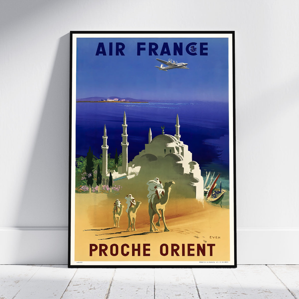 A framed vintage-style Air France travel poster leaning against a white wall on a light floor. The poster shows an airplane over a desert scene with camels, a domed building, and the text 'AIR FRANCE' at the top and 'PROCHE ORIENT' at the bottom.