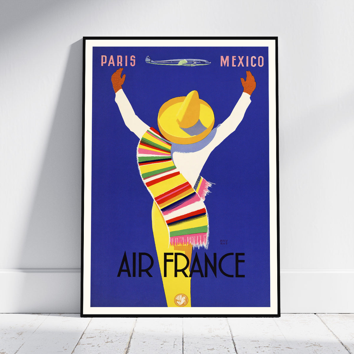 A framed vintage-style travel poster leaning against a white wall on a light floor. The poster shows a stylized figure in a yellow hat and multicolored striped wrap on a blue background, with the words PARIS, MEXICO, AIR FRANCE, and a small airplane illustration.