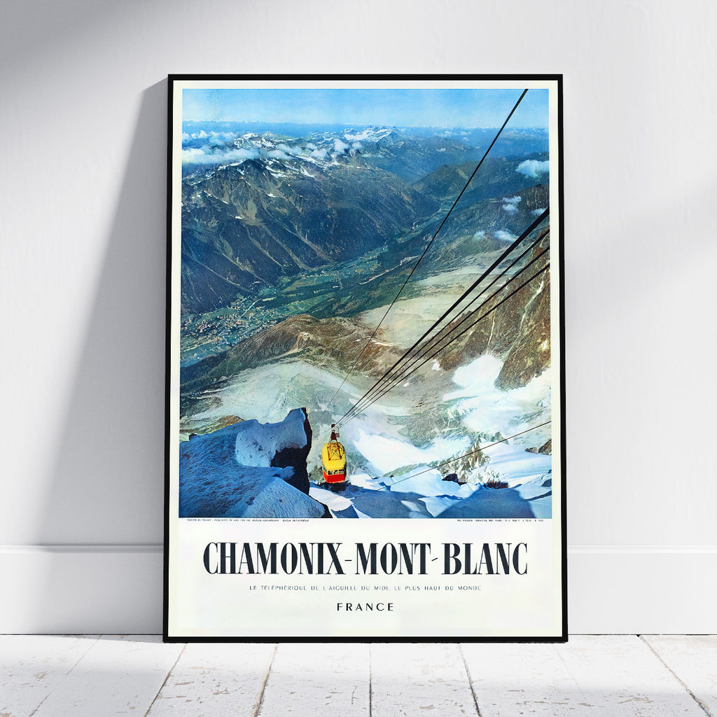 A framed vintage-style travel poster leaning against a white wall on a light floor. The poster shows a mountain valley with snow, a gondola, and the text 'CHAMONIX-MONT BLANC' and 'FRANCE'.