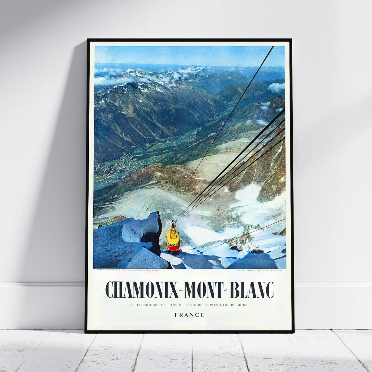 A framed vintage-style travel poster leaning against a white wall on a light floor. The poster shows a mountain valley with snow, a gondola, and the text 'CHAMONIX-MONT BLANC' and 'FRANCE'.