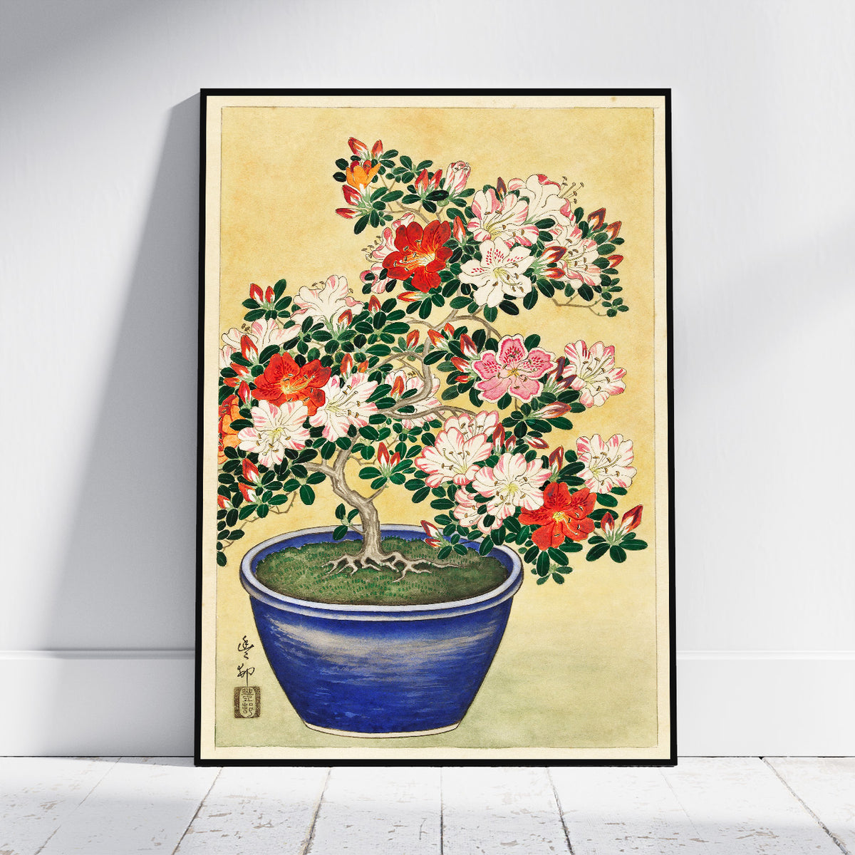 A framed art print depicting a traditional Japanese-style bonsai tree with red, pink, and white flowers in a blue pot, leaning against a white wall on a light wood floor.