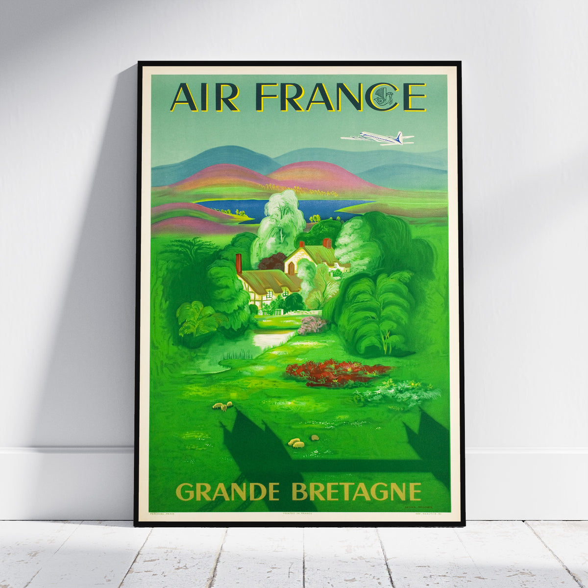 A framed vintage-style Air France travel poster leaning against a white wall on a light wood floor. The poster features a green landscape with rolling hills, trees, a small house, and an airplane, with the text 'AIR FRANCE' at the top and 'GRANDE BRETAGNE' at the bottom.