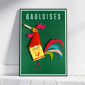 A framed vintage-style poster leaning against a white wall on a light wood floor. The poster features the word 'GAULOISES' at the top and a stylized rooster holding a Gaulois bottle, with a green background and colorful feathers.