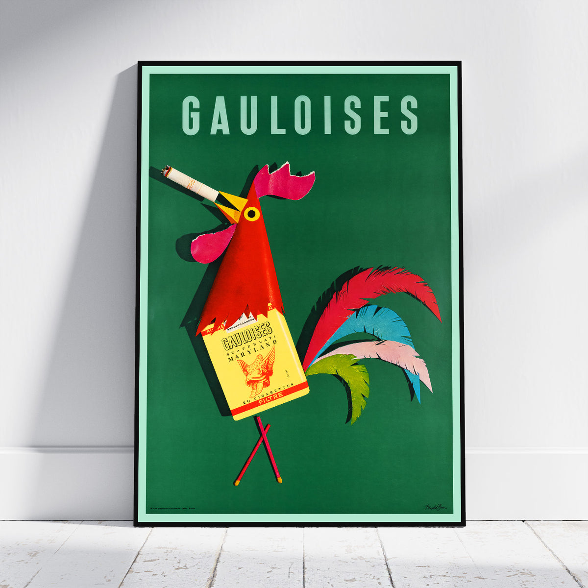 A framed vintage-style poster leaning against a white wall on a light wood floor. The poster features the word 'GAULOISES' at the top and a stylized rooster holding a Gaulois bottle, with a green background and colorful feathers.