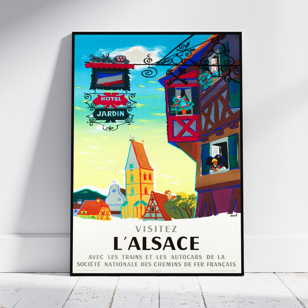 A framed travel poster for Alsace, France, leaning against a white wall on a light wood floor. The poster features a colorful illustration of a village with a tower, a red-roofed house, and a sign reading 'HOTEL JARDIN' and 'VISITEZ L'ALSACE'.