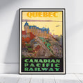 A framed vintage-style travel poster for Quebec and the Canadian Pacific Railway, leaning against a white wall on a light floor. The poster features a yellow sky, a large castle, colorful buildings, and green lettering at the bottom.