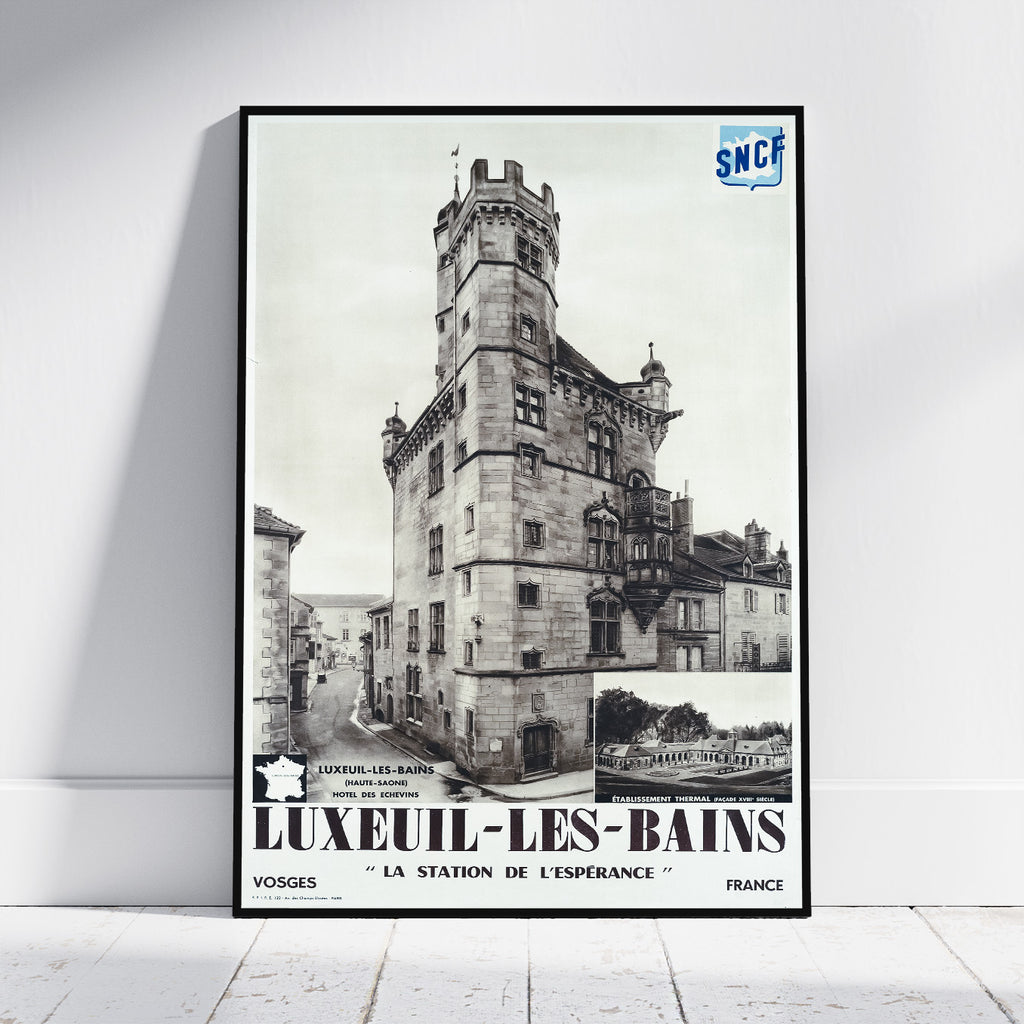 A framed vintage travel poster for Luxeuil-Les-Bains, France, leaning against a white wall on a light wood floor. The poster features a black and white photograph of a historic stone tower and station building, with bold black text at the bottom and a blue SNCF logo in the top right corner.