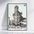 A framed vintage travel poster for Luxeuil-Les-Bains, France, leaning against a white wall on a light wood floor. The poster features a black and white photograph of a historic stone tower and station building, with bold black text at the bottom and a blue SNCF logo in the top right corner.