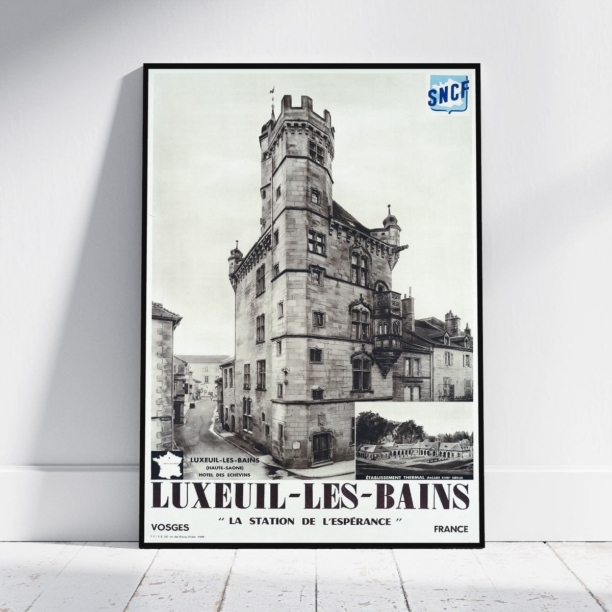A framed vintage travel poster for Luxeuil-Les-Bains, France, leaning against a white wall on a light wood floor. The poster features a black and white photograph of a historic stone tower and station building, with bold black text at the bottom and a blue SNCF logo in the top right corner.