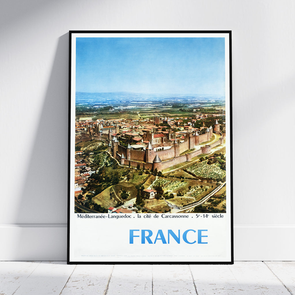 A framed vintage-style travel poster of Carcassonne, France, leaning against a white wall on a light wood floor. The poster features a black frame, a white border, and a colorful aerial illustration of the historic city under a blue sky, with 'FRANCE' printed in large blue letters at the bottom.