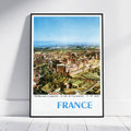 A framed vintage-style travel poster of Carcassonne, France, leaning against a white wall on a light wood floor. The poster features a black frame, a white border, and a colorful aerial illustration of the historic city under a blue sky, with 'FRANCE' printed in large blue letters at the bottom.