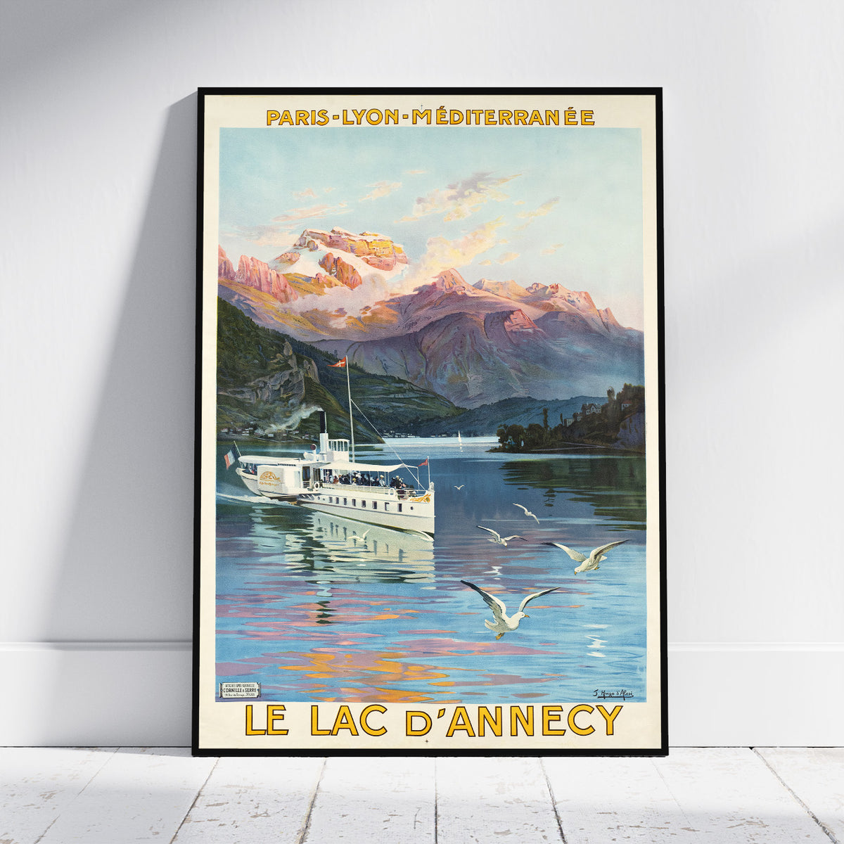 A framed vintage-style travel poster leaning against a white wall on a light wood floor. The artwork shows a boat on a blue lake with mountains, seagulls, and the text 'PARIS-LYON-MÉDITERRANÉE' at the top and 'LE LAC D'ANNECY' at the bottom.