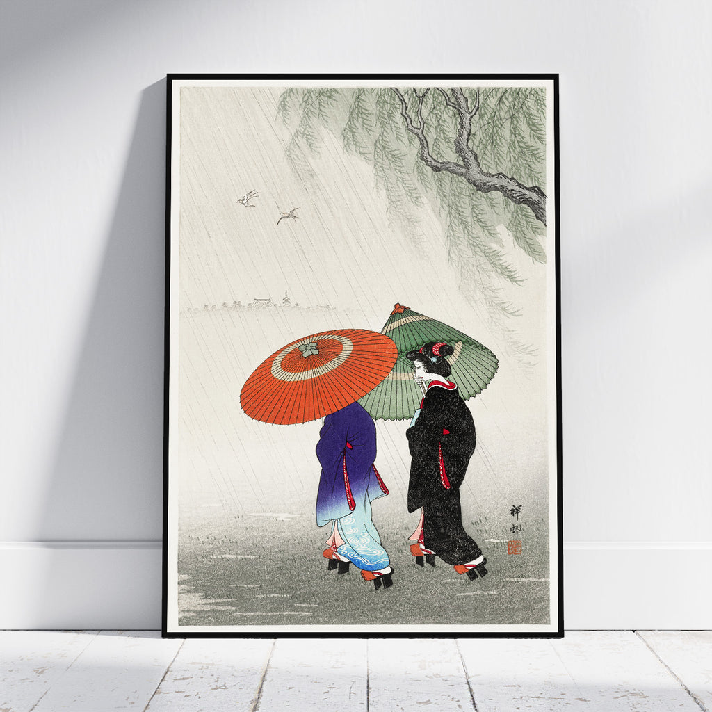 A framed art print leaning against a white wall on a light wood floor. The print features a Japanese-style illustration of two women walking in the rain under a weeping willow tree, one holding a bright orange umbrella and the other a green umbrella.
