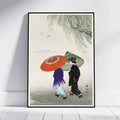 A framed art print leaning against a white wall on a light wood floor. The print features a Japanese-style illustration of two women walking in the rain under a weeping willow tree, one holding a bright orange umbrella and the other a green umbrella.