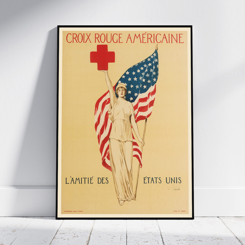 A framed vintage-style American Red Cross poster leaning against a white wall on a light wood floor. The poster shows a woman holding an American flag with a red cross above her head, with the French text 'CROIX ROUGE AMÉRICaine' at the top and 'L’AMITIÉ DES ÉTATS UNIS' at the bottom.