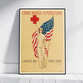 A framed vintage-style American Red Cross poster leaning against a white wall on a light wood floor. The poster shows a woman holding an American flag with a red cross above her head, with the French text 'CROIX ROUGE AMÉRICaine' at the top and 'L’AMITIÉ DES ÉTATS UNIS' at the bottom.