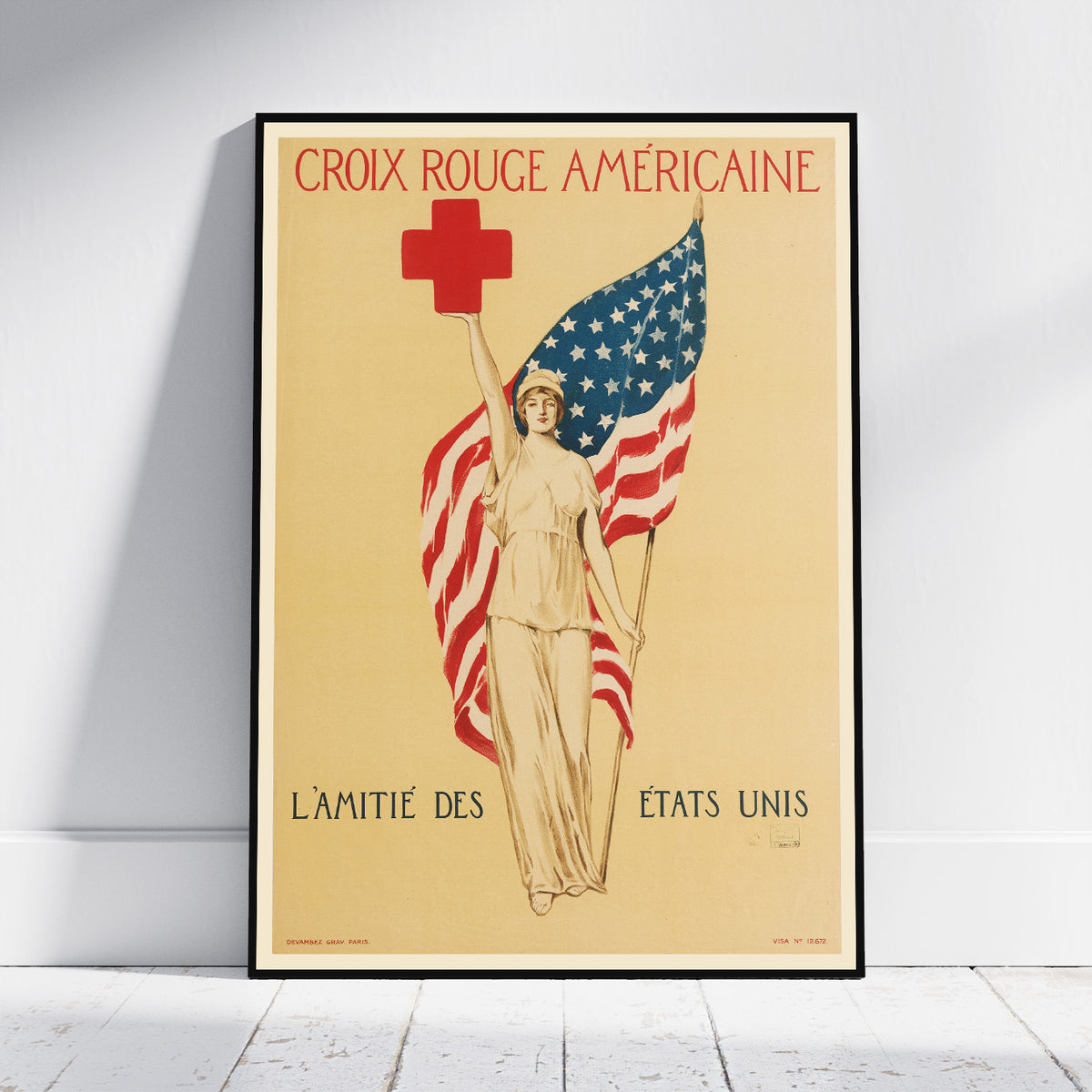A framed vintage-style American Red Cross poster leaning against a white wall on a light wood floor. The poster shows a woman holding an American flag with a red cross above her head, with the French text 'CROIX ROUGE AMÉRICaine' at the top and 'L’AMITIÉ DES ÉTATS UNIS' at the bottom.