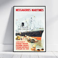 A framed vintage-style shipping poster leaning against a white wall on a light wood floor. The poster features the text 'MESSAGERIES MARITIMES' in red at the top and additional red text near the bottom, with an illustration of a large ocean liner docked at a harbor and small boats in the foreground.