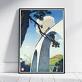 A framed vintage-style travel poster leaning against a white wall on a light wood floor. The artwork shows a large white arched bridge over a blue sky, with pine branches across the top, distant mountains, and green hills.
