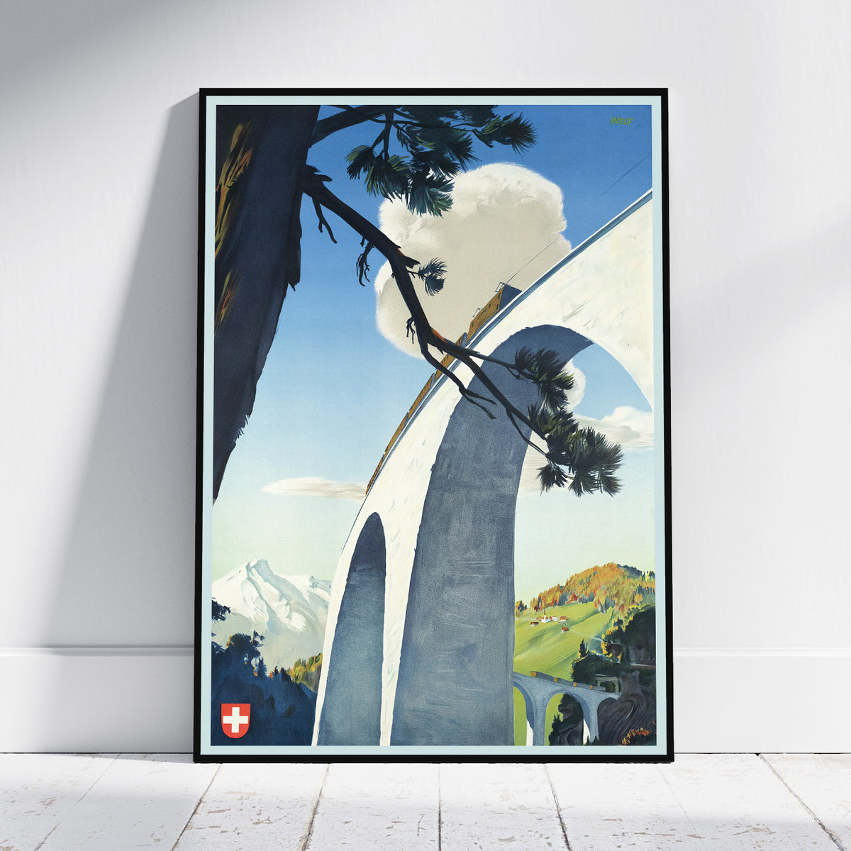 A framed vintage-style travel poster leaning against a white wall on a light wood floor. The artwork shows a large white arched bridge over a blue sky, with pine branches across the top, distant mountains, and green hills.
