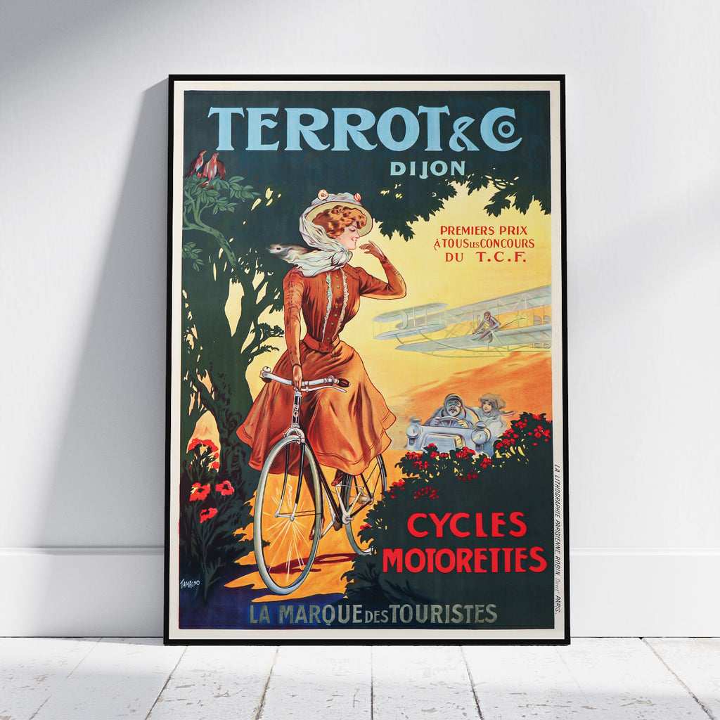 A framed vintage poster print leaning against a white wall on a light wood floor. The poster features an illustrated woman riding a bicycle in a terracotta dress, with the text 'TERROT & C DIJON' at the top and 'CYCLES MOIROTTES' at the bottom.