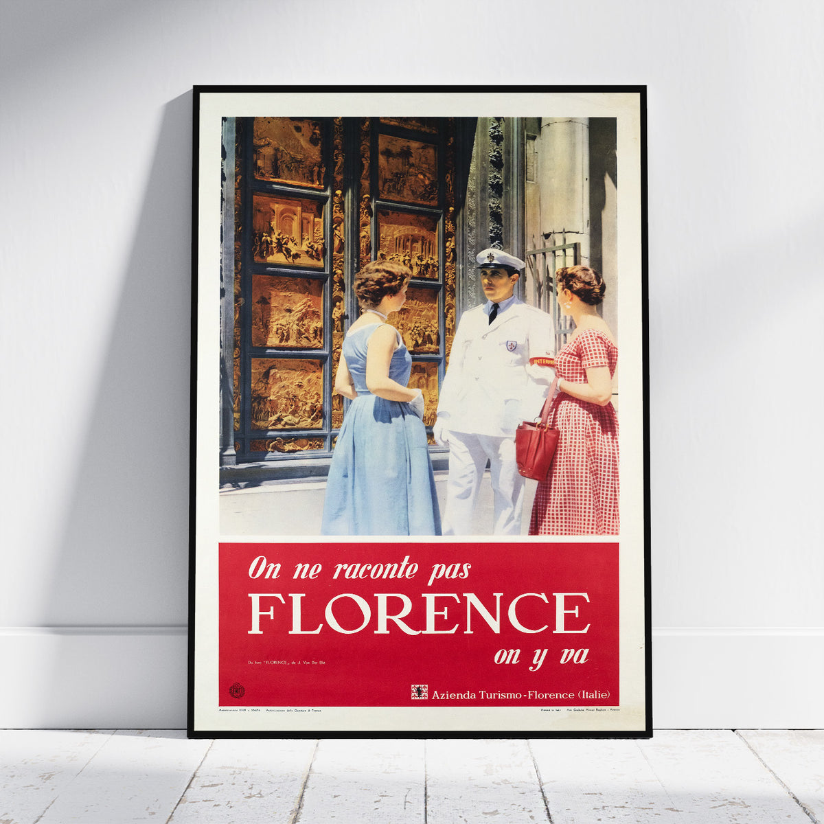 A framed vintage travel poster leaning against a white wall on a light wood floor. The poster features a black-and-white photograph of three people in front of ornate doors, with a bold red lower section containing white Spanish text reading 'On no raconto pas FLORENCE on y pa.