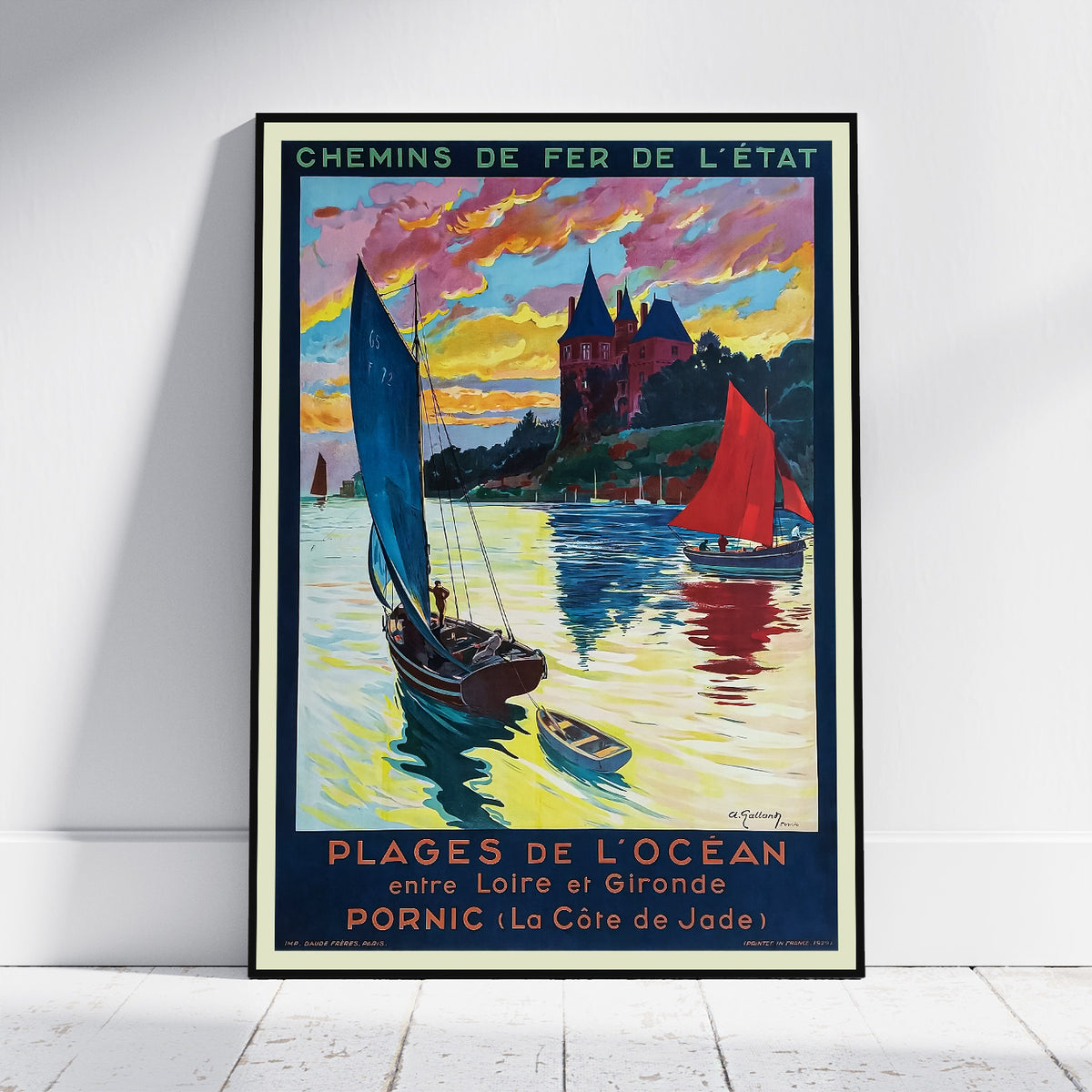 A framed vintage-style travel poster leaning against a white wall on a light wood floor. The poster depicts colorful sailboats on water with a castle and dramatic sunset sky.