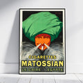 A framed vintage-style poster leaning against a white wall on a light floor. The poster shows a person wearing a large green turban and smoking, with bold yellow text reading 'CIGARETTES MATOSSIAN LE CAIRE ÉGYPTE'.