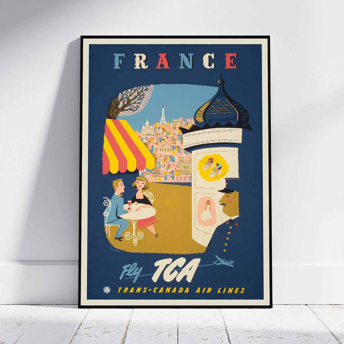 A framed vintage-style travel poster leaning against a white wall on a light floor. The poster reads "FRANCE" at the top and "Fly TCA Trans-Canada Air Lines" at the bottom, with illustrated elements including a city skyline, a striped awning, café scene, and a tower.
