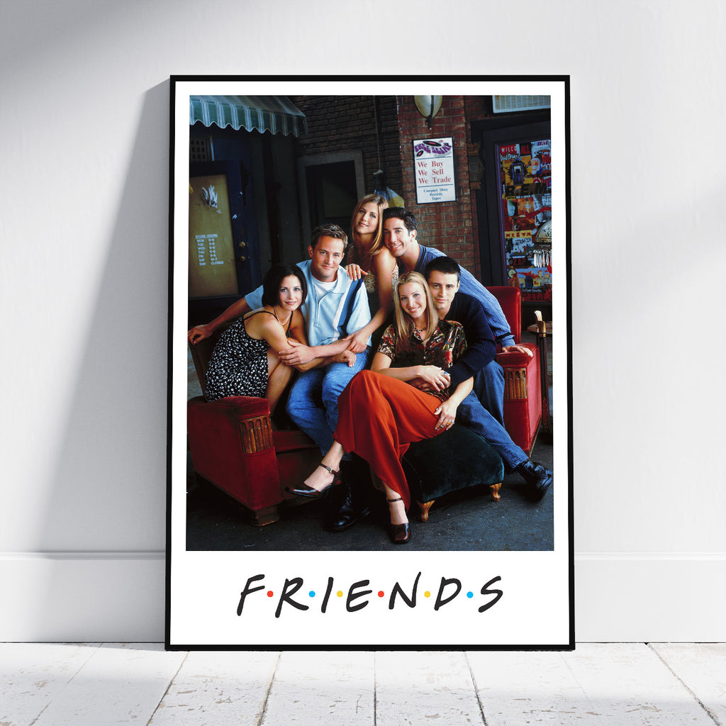 A framed Friends poster leaning against a white wall on a light wood floor. The poster features a black-framed photo of the iconic cast of Friends sitting on a red couch, with the 'FRIENDS' logo printed below in the classic multicolor dot style.