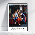 A framed Friends poster leaning against a white wall on a light wood floor. The poster features a black-framed photo of the iconic cast of Friends sitting on a red couch, with the 'FRIENDS' logo printed below in the classic multicolor dot style.