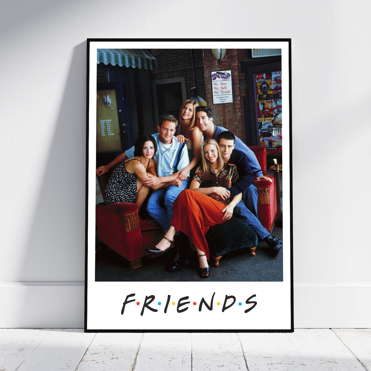 A framed Friends poster leaning against a white wall on a light wood floor. The poster features a black-framed photo of the iconic cast of Friends sitting on a red couch, with the 'FRIENDS' logo printed below in the classic multicolor dot style.
