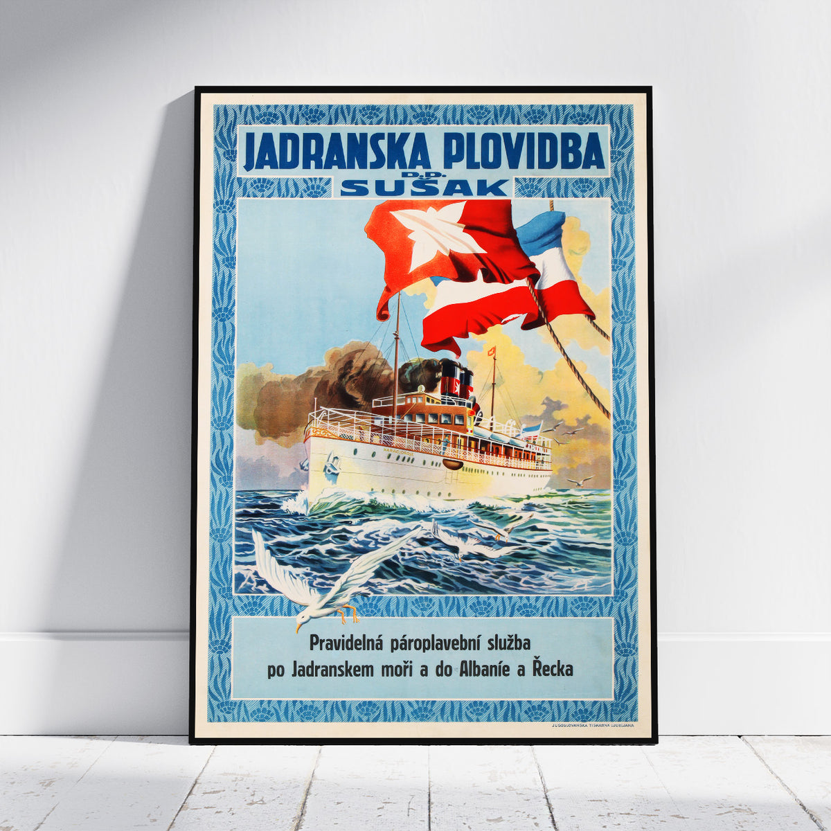 A framed vintage-style travel poster leaning against a white wall on a light wood floor. The poster features a white passenger ship on blue ocean waves with red and blue flags, and the text 'JADRANSKA PLOVIDBA SUŠAK' at the top.