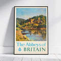 A framed vintage-style travel poster leaning against a white wall on a light wood floor. The poster shows a scenic landscape with a river, green hills, and an abbey, with the text 'The Abbeys of BRITAIN' at the bottom.
