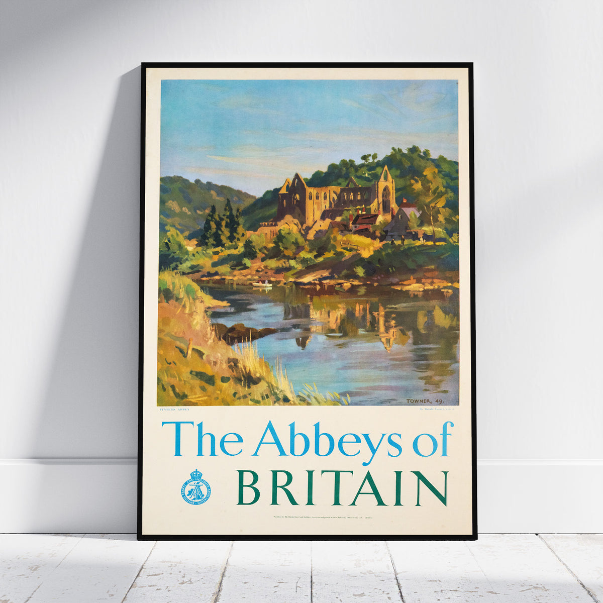 A framed vintage-style travel poster leaning against a white wall on a light wood floor. The poster shows a scenic landscape with a river, green hills, and an abbey, with the text 'The Abbeys of BRITAIN' at the bottom.
