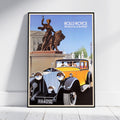 A framed vintage Rolls-Royce poster leaning against a white wall on a light floor. The poster shows an illustration of a yellow classic car in the foreground and a bronze equestrian statue in the background, with the text 'ROLLS ROYCE The Best Car in the World' at the top.