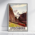A framed vintage-style travel poster of the Lötschberg mountain train in Switzerland, leaning against a white wall on a light wood floor. The poster depicts a red train traveling through a scenic alpine valley with rocky cliffs and snow-capped mountains under a pale sky.