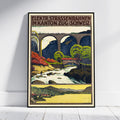 A framed vintage-style travel poster leaning against a white wall on a light floor. The poster shows an illustrated mountain railway scene with arches, trees, a river, and bold text at the top reading 'ELEKTR.