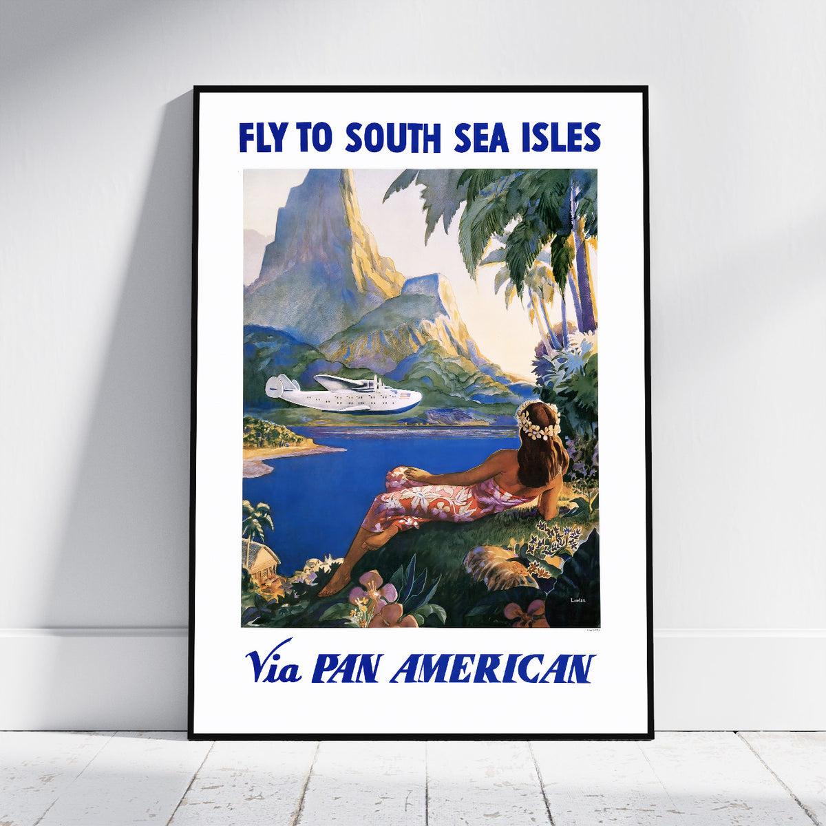 A framed vintage-style travel poster leaning against a white wall on a light floor. The poster reads 'FLY TO SOUTH SEA ISLES' at the top and 'Via PAN AMERICAN' at the bottom, with an illustrated tropical scene showing blue water, mountains, palm trees, and a woman reclining by the shore.