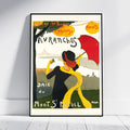 A framed vintage-style travel poster leaning against a white wall on a light wood floor. The poster features an Art Deco illustration of a woman in a black dress and red hat holding a red umbrella, with a yellow scarf, green landscape, and French text including 'Avranches' and 'Mont Saint-Michel'.