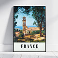 A vertical framed travel poster of a French coastal village with a church tower, trees, and the word FRANCE at the bottom, leaning against a white wall on a light floor.