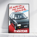 A framed vintage-style Mazda poster leaning against a white wall on a light floor. The poster shows a black Mazda car with red French text reading 'Et avec ça, un punch terrible.