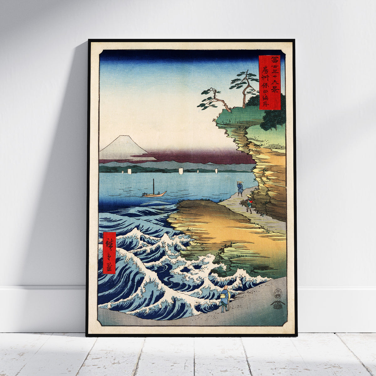 A framed Japanese-style art print leaning against a white wall on a light floor. The artwork depicts blue ocean waves, a sandy shore, cliffs, a tree, distant mountains, and red vertical label elements with Japanese text.