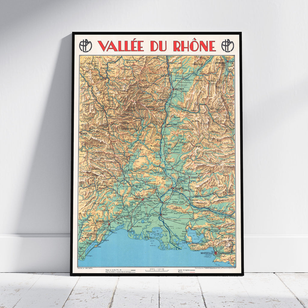 A framed vintage-style topographic map poster of the Vallée du Rhône leaning against a white wall on a light wood floor. The poster features a black frame, red title text at the top, and a multicolored map with beige, green, blue, and brown details.