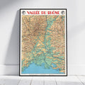 A framed vintage-style topographic map poster of the Vallée du Rhône leaning against a white wall on a light wood floor. The poster features a black frame, red title text at the top, and a multicolored map with beige, green, blue, and brown details.