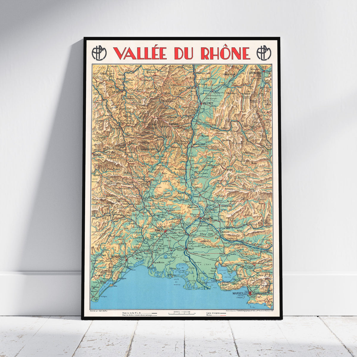 A framed vintage-style topographic map poster of the Vallée du Rhône leaning against a white wall on a light wood floor. The poster features a black frame, red title text at the top, and a multicolored map with beige, green, blue, and brown details.