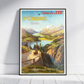 A framed vintage-style travel poster leaning against a white wall on a light floor. The poster depicts a scenic mountain landscape with a lake, green hills, and a road with horse-drawn and carriages.