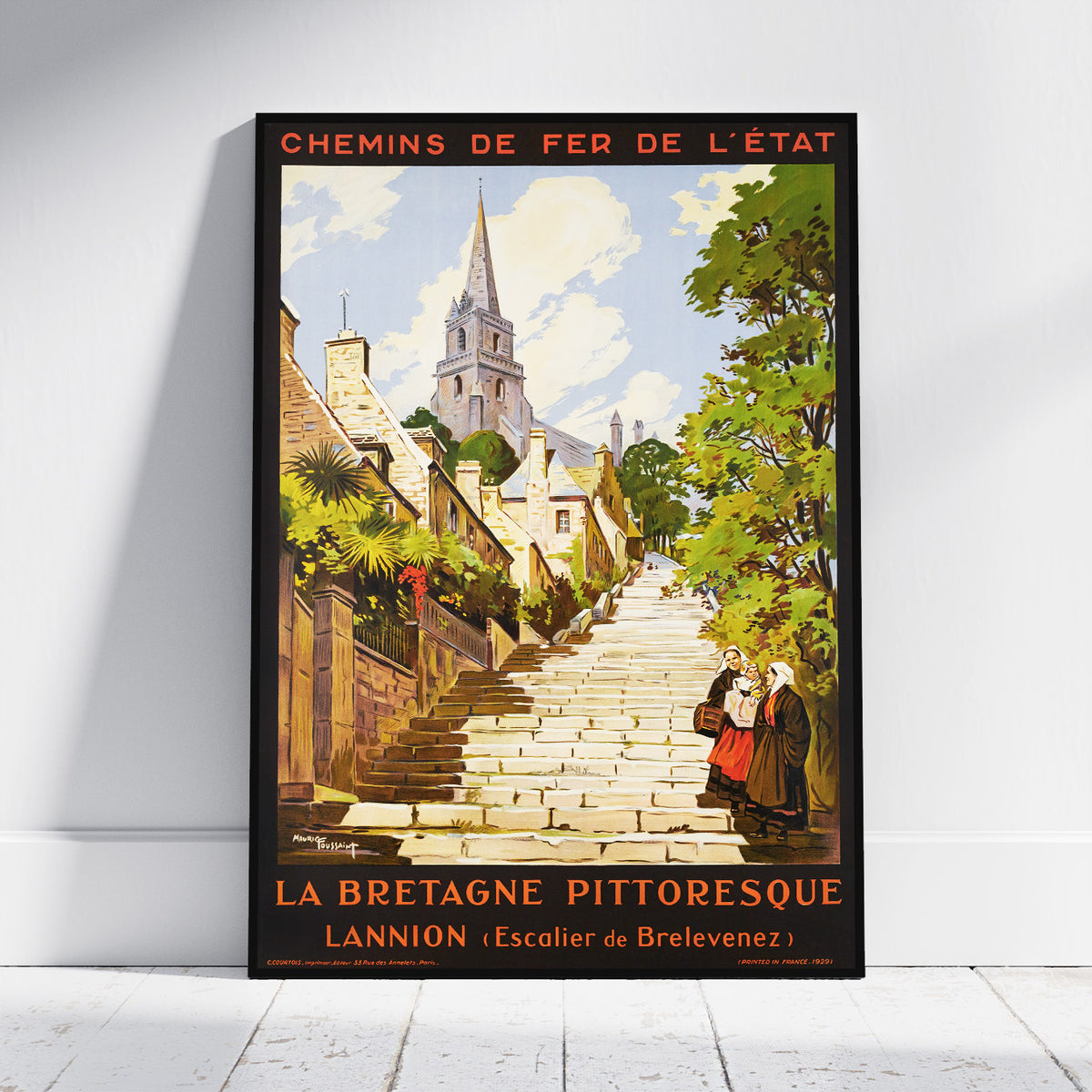 A framed vintage-style travel poster leaning against a white wall on a light floor. The poster shows a stone staircase leading upward through a scenic town with trees, buildings, and a church spire, with French text at the top and bottom.