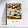 A framed vintage-style travel poster of the Roman arena in Nîmes, France, leaning against a white wall on a light wood floor. The poster features an aerial view of the arena and surrounding city, with 'FRANCE' printed in large orange letters at the bottom.