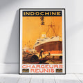 A framed vintage-style travel poster leaning against a white wall on a light floor. The poster reads "INDOCHINE" at the top and "CHARGEURS REUNIS" at the bottom, with an illustration of a large ship at a harbor in warm orange, yellow, and brown tones.